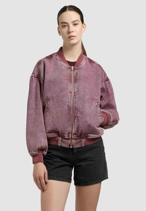 Moteris su tamsiais plaukais, surištais į uodegą, dėvi išblukusią violetinę bombonešio striukę ir juodus džinso šortus, stovinti priešais paprastą šviesų foną.