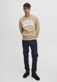 Beige mikina s nápisem "CASUAL FRIDAY" v tučném bílém písmu, v kombinaci s tmavě modrými džíny a černými botami. Minimalistický design a pohodlný střih.