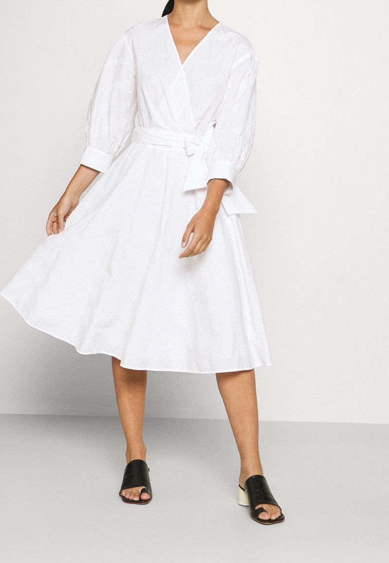 Robe blanche à manches bouffantes, taille cintrée et jupe évasée. Caractéristiques d'un tissu texturé avec des motifs subtils. Associée à des sandales noires.