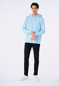 Felpa leggera blu con tasca frontale e cappuccio con lacci, presenta testo bianco. Abbinata a jeans neri e sneakers bianche.