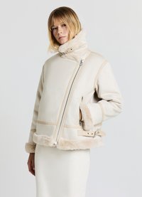 Giacca crema foderata in shearling con doppia zip, colletto morbido e dettagli con cintura; presenta una superficie esterna liscia e cuciture a contrasto.