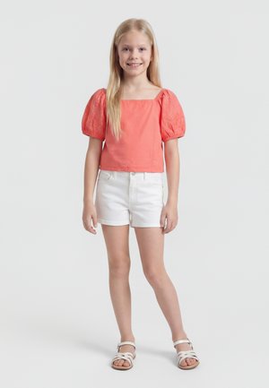 Fiatal lány hosszú szőke hajjal, korall színű puffos ujjú felsőt, fehér rövidnadrágot és fehér szandált visel, miközben áll és mosolyog egyszínű háttér előtt.