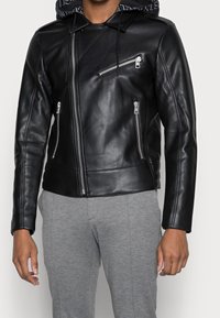 Giacca da moto nera in pelle con cappuccio, dotata di zip in argento e vestibilità slim. La giacca ha una texture liscia e un design moderno.