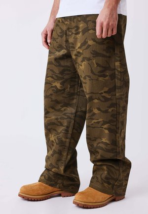 Homme portant un pantalon camouflage ample vert et marron ainsi que des bottes de travail beige, debout sur un fond blanc.