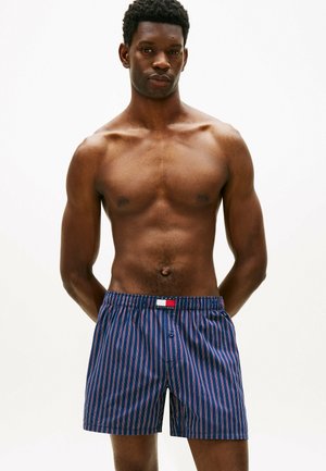 Man zonder shirt, gekleed in blauw gestreepte boxershorts met een Tommy Hilfiger-logo, staand met de handen achter zijn rug tegen een effen achtergrond.