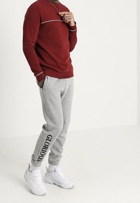 Sudadera roja con un diseño de rayas blancas combinada con pantalones de chándal grises que llevan "GLORIOUS" en negro a lo largo del costado. Zapatillas blancas completan el conjunto.