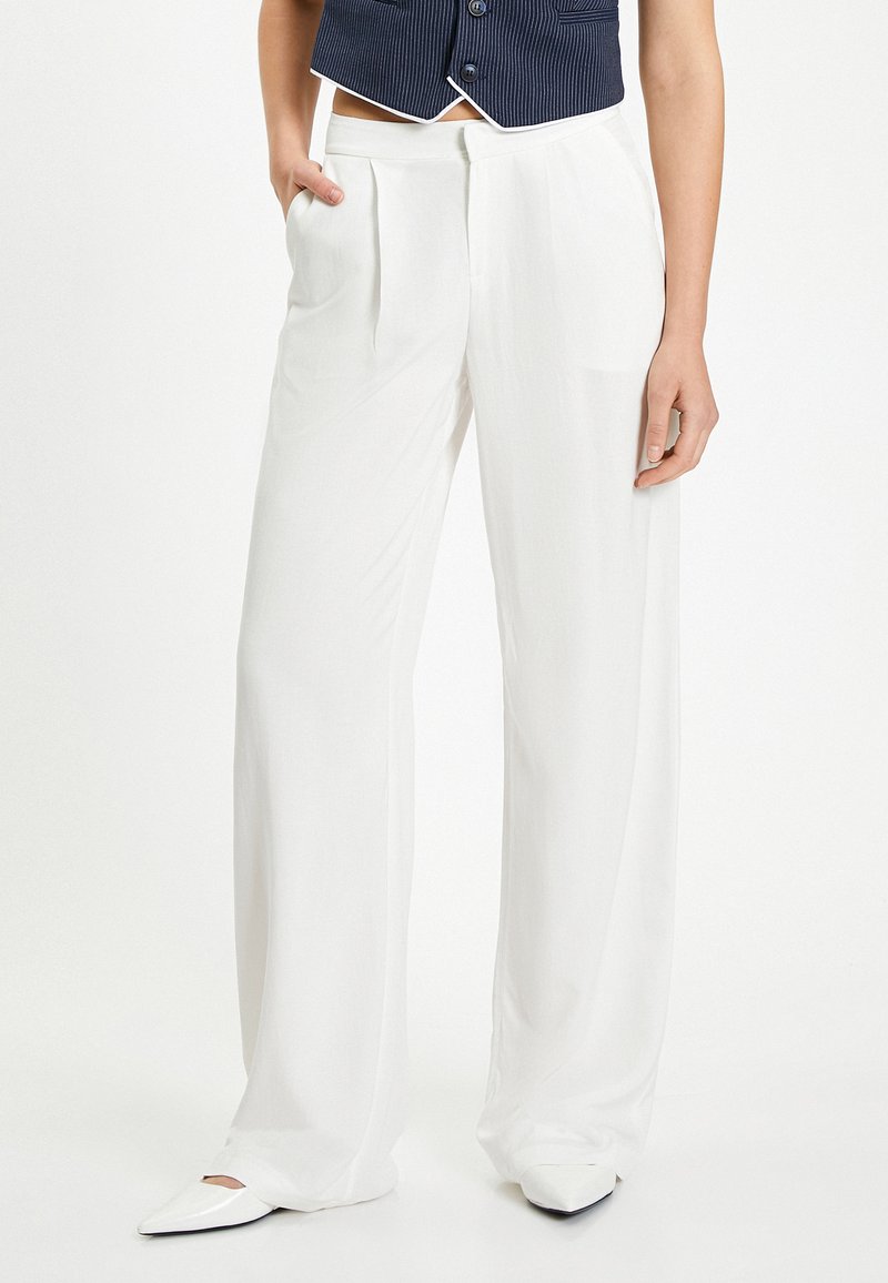 Pantalons larges blancs avec une texture lisse, dotés de plis sur le devant et de poches latérales. Associés à des chaussures blanches pointues et un gilet navy.
