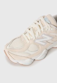 Baskets New Balance beiges et blanches avec tissu en maille, superpositions en daim, semelle épaisse et lacets sur fond blanc.