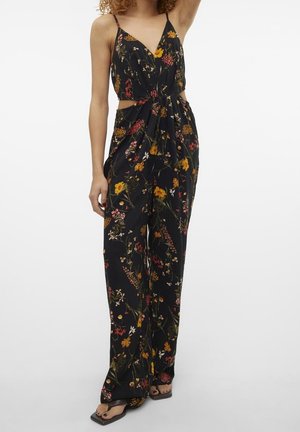 Kobieta ubrana w czarny kombinezon w kwiaty z cienkimi ramiączkami, wycięciami po bokach, szerokimi nogawkami i czarne sandały na kwadratowym nosku.