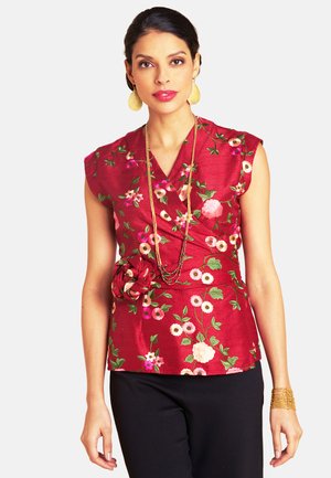 Rotes, mit Blumen besticktes Oberteil mit V-Ausschnitt, kurzen Kappärmeln und seitlichem Blumendetail, kombiniert mit schwarzer Hose und geschichteten Halsketten.