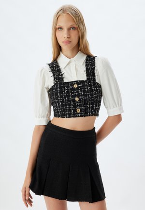 Top corto a quadratini in bianco e nero con bottoni e spalline sfrangiate, abbinato a una gonna plissettata nera con texture. Entrambi hanno una vestibilità tailored.