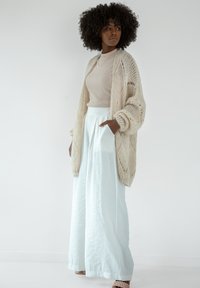 Jasno beżowy sweter z dzianiny z oversize'owymi rękawami, połączony z jasnoniebieską spódnicą o szerokich nogawkach. Modelka stoi na tle białym.