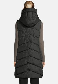 Woman with blond ponytail wearing a long black quilted sleeveless puffer vest with a high collar, viewed from the back.