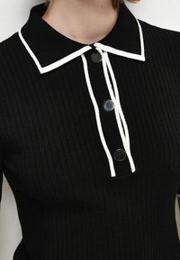 Haut en maille côtelée noire avec col blanc à revers et boutons contrastants. Présente une coupe ajustée et un détail décoratif noué.