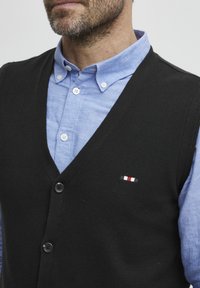 Svart ullväst med V-ringning, prydd med två knappar och en liten broderad logotyp i röd, vit och svart på bröstet. Ljusblå skjorta under.