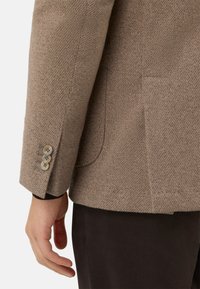 Blazer in lana marrone con motivo a spina di pesce, bavero con tacca, dettaglio a tre bottoni sui polsini e tasca laterale, abbinato a pantaloni in tessuto scuro.