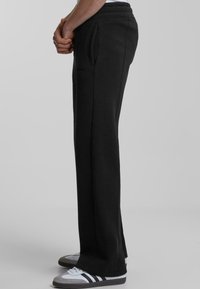 Black, relaxed-fit sweatpants with a drawstring waistband, side pockets, and a smooth texture. Featured with white and gray sneakers.
