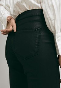 Personne portant un pantalon noir et une chemise blanche à manches longues, avec une main posée près de la poche arrière du pantalon.
