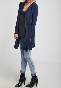 Navy knitted cardigan with a textured pattern, open front, and long sleeves, worn over a black top and paired with light blue jeans and black ankle boots.