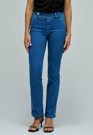 Mujer con vaqueros azules de talle alto con botones laterales delanteros y sandalias negras de tacón, de pie sobre fondo liso.