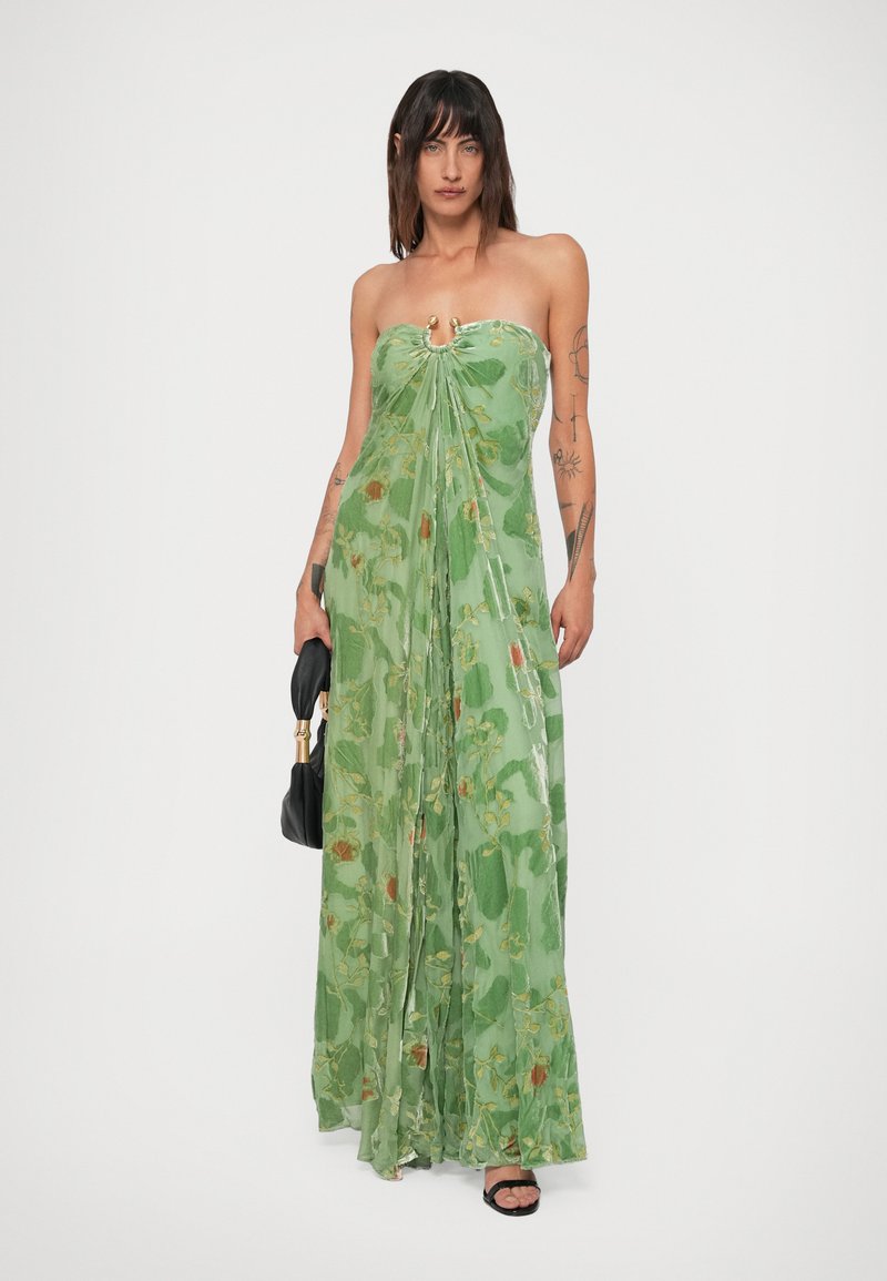 Woman with tattoos wears strapless green floral maxi dress, holding black handbag, standing against white background.