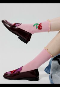 Pair of legs with pink socks featuring green and red palm tree design, wearing shiny dark burgundy loafers with tassels.