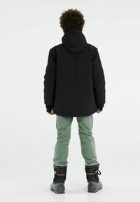 Schwarze wasserdichte Jacke mit Kapuze, enganliegenden Bündchen und lässiger Passform, kombiniert mit grünen Hosen und schwarzen isolierten Stiefeln.