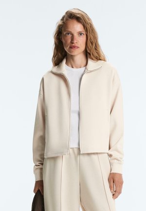 Frau mit hellen, welligen Haaren trägt eine beige Reißverschlussjacke über einem weißen Hemd und passenden beigen Hosen, sie hält ein dunkles Objekt in ihrer linken Hand.