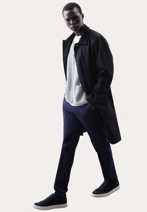 Mand går, iført sort lang frakke, grå lynlåstrøje, navy bukser og sorte sneakers med hvide såler.