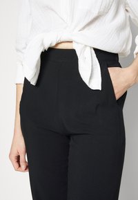 Svarta skräddarsydda byxor med en slät textur, midjehög midja och sidofickor, kompletterade med en vit, strukturerad knäpptröja knuten vid midjan.