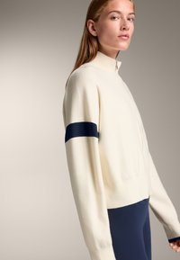 Cremefarbener Zip-Up-Pullover mit hohem Kragen, ausgestattet mit einem strukturierten marineblauen Streifen über dem Ärmel. Weicher, gestrickter Stoff, lockere Passform.