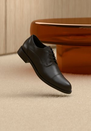 Fekete bőrből készült, fűzős, elegáns oxford cipő lebegő hatással bézs szőnyeg felett, barna kerek asztal látható a háttérben.