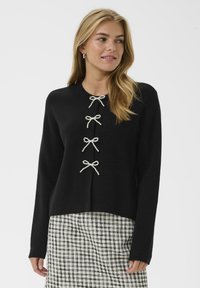 Svart stickad cardigan med fem fuskpärlknappar i form av rosetter fram. Långa ärmar och rundad halsringning. Matchad med en mönstrad kjol.