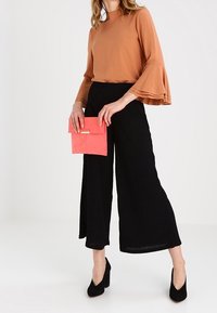 Jasnobrązowa, długosleevowa bluzka z szerokimi rękawami; czarne spodnie o szerokich nogawkach; trzyma różową kopertówkę; czarne szpilki z ostrym noskiem.