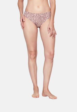 Frau trägt hellbraune Bikinihöschen mit Leopardenmuster, steht barfuß vor einem schlichten weißen Hintergrund.