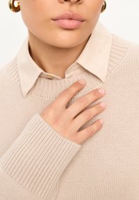 Lichtbeige gebreide trui met geribde mouwen, gelaagd over een crèmekleurig overhemd met kraag. Glad textuur en casual ontwerp, met een warme kleurenpalet.