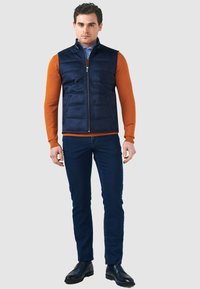 Marineblaue gefütterte Weste mit hohem Kragen, Frontreißverschluss und zwei Seitentaschen, kombiniert mit einem orangefarbenen Langarmshirt und dunkler Jeans.