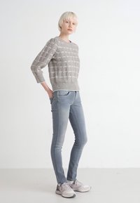 Pull en maille gris clair avec un motif texturé, associé à un jean bleu clair ajusté. Modèle portant des chaussures de sport, posé les mains dans les poches.