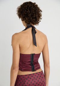 Burgundy polka dot corset top with a halter neck, black ribbon tie, and a visible zip closure at the back. Smooth satin fabric texture.