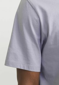 Camisa de manga corta en algodón lavanda claro, con cuello redondo, textura suave y costuras sutiles a lo largo del dobladillo y las mangas.