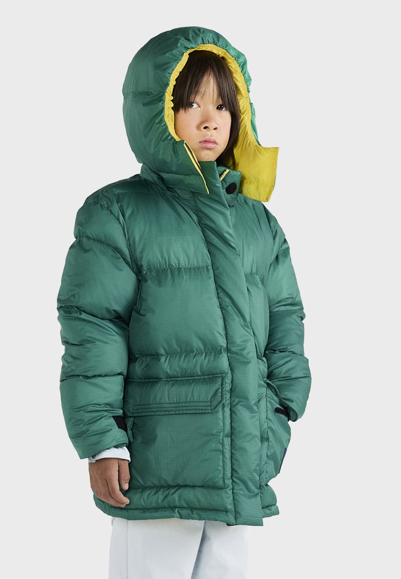 Chaqueta acolchada verde con capucha, que presenta un forro amarillo en contraste, bolsillos frontales y un diseño acolchado. Texturizada, cálida e isolante.