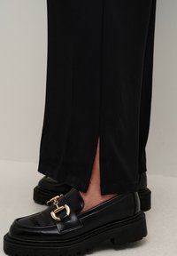 Černé loafers s detailem zlaté spony, nošené s černými kalhotami s malým bočním rozparkem nad kotníkem.