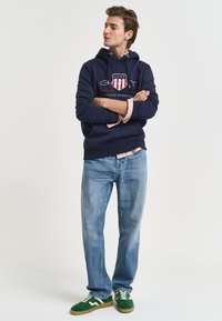 Felpa navy con logo e dettagli rossi, camicia chiara sotto, jeans azzurri chiari, sneaker verdi con dettagli bianchi, in piedi con le braccia incrociate.