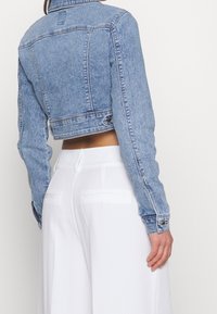 Osoba oblečená v krátké modré džínové bundě a vysokým pasem bílých kalhot, zobrazená zezadu na neutrálním pozadí.