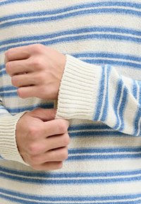 Mains ajustant le poignet d'un pull tricoté crème et bleu à rayures, avec une texture côtelée sur les manches et le corps.