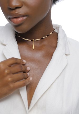 Nahaufnahme einer Person, die ein weißes Hemd und ein dünnes, leopardgemustertes Halsband mit einem kleinen goldenen Anhänger trägt, die Hand berührt den Kragen.