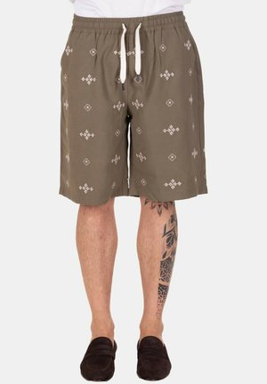 Uomo che indossa pantaloncini verde oliva con motivi geometrici bianchi, una coulisse bianca, scarpe slip-on nere e un grande tatuaggio nero sul polpaccio destro.