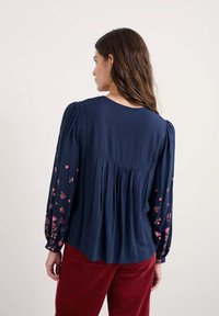 Blusa blu navy con maniche a sbuffo e ricami floreali in rosa e rosso. Presenta un design con schiena plissettata, texture morbida e collo rotondo.