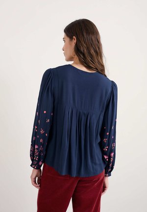 Navyblauwe blouse met pofmouwen en bloemenborduursel in roze en rood. Heeft een geplooid achterontwerp, zachte textuur en ronde hals.