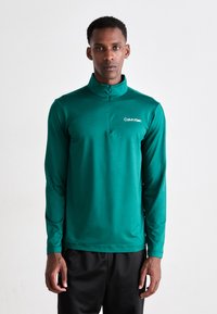 Pullover verde a maniche lunghe con mezza zip e colletto alto. Presenta il logo di Calvin Klein in bianco sul lato sinistro del petto. Tessuto morbido e leggero.